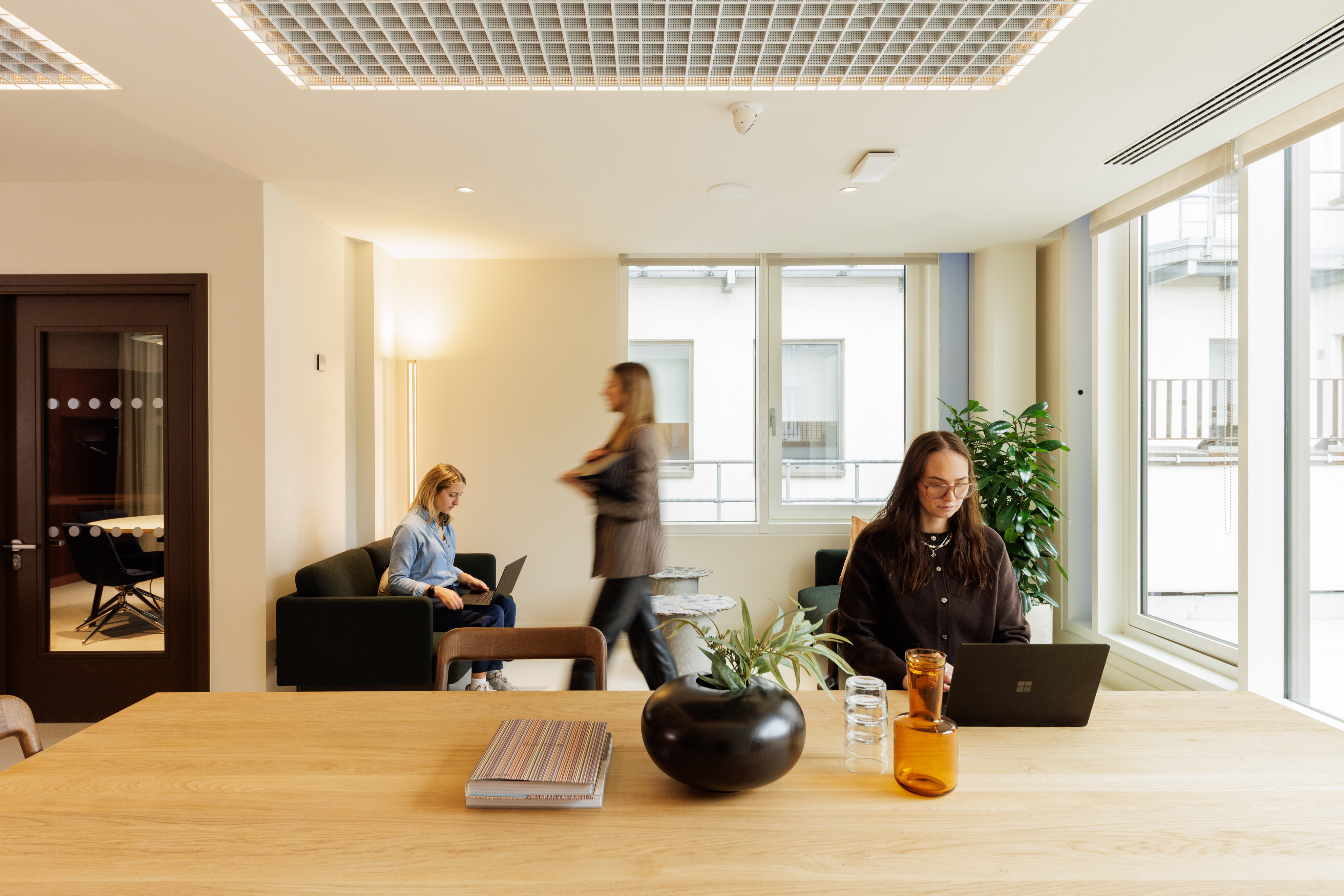 Professionals working and moving through a light-filled communal workspace with lounge seating and collaborative areas.