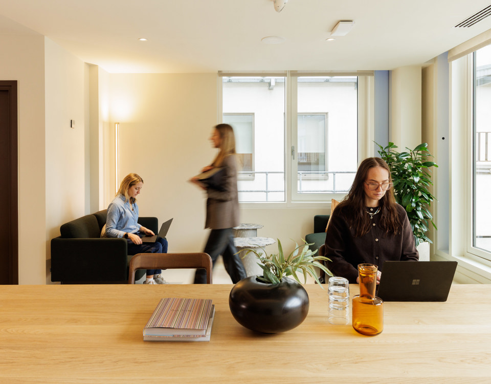 Professionals working and moving through a light-filled communal workspace with lounge seating and collaborative areas.