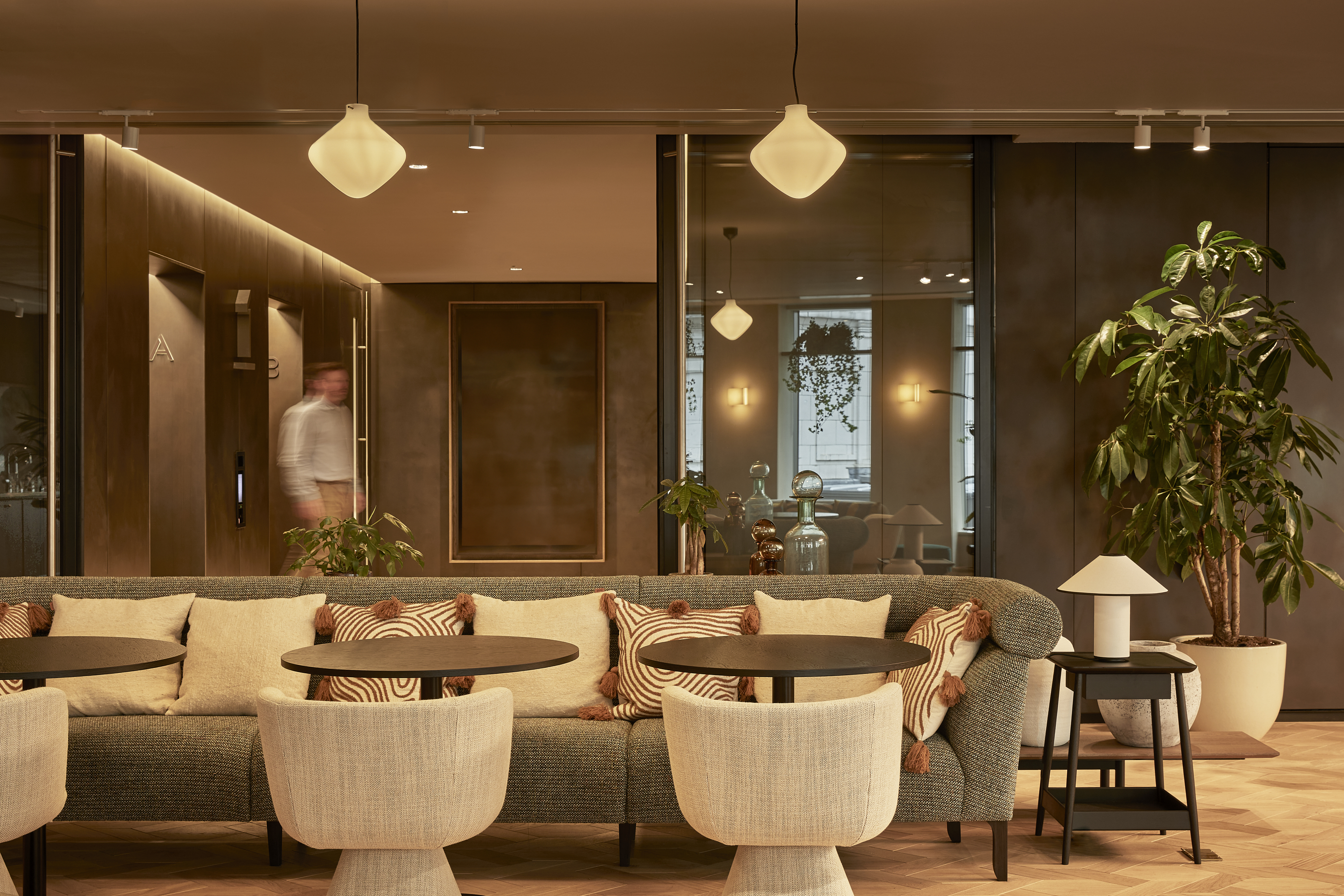 A contemporary lounge area featuring a large sofa, upholstered chairs, side tables, indoor plants and glass-fronted meeting rooms in a modern workspace.