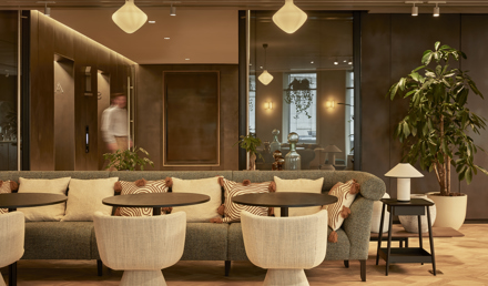 A contemporary lounge area featuring a large sofa, upholstered chairs, side tables, indoor plants and glass-fronted meeting rooms in a modern workspace.