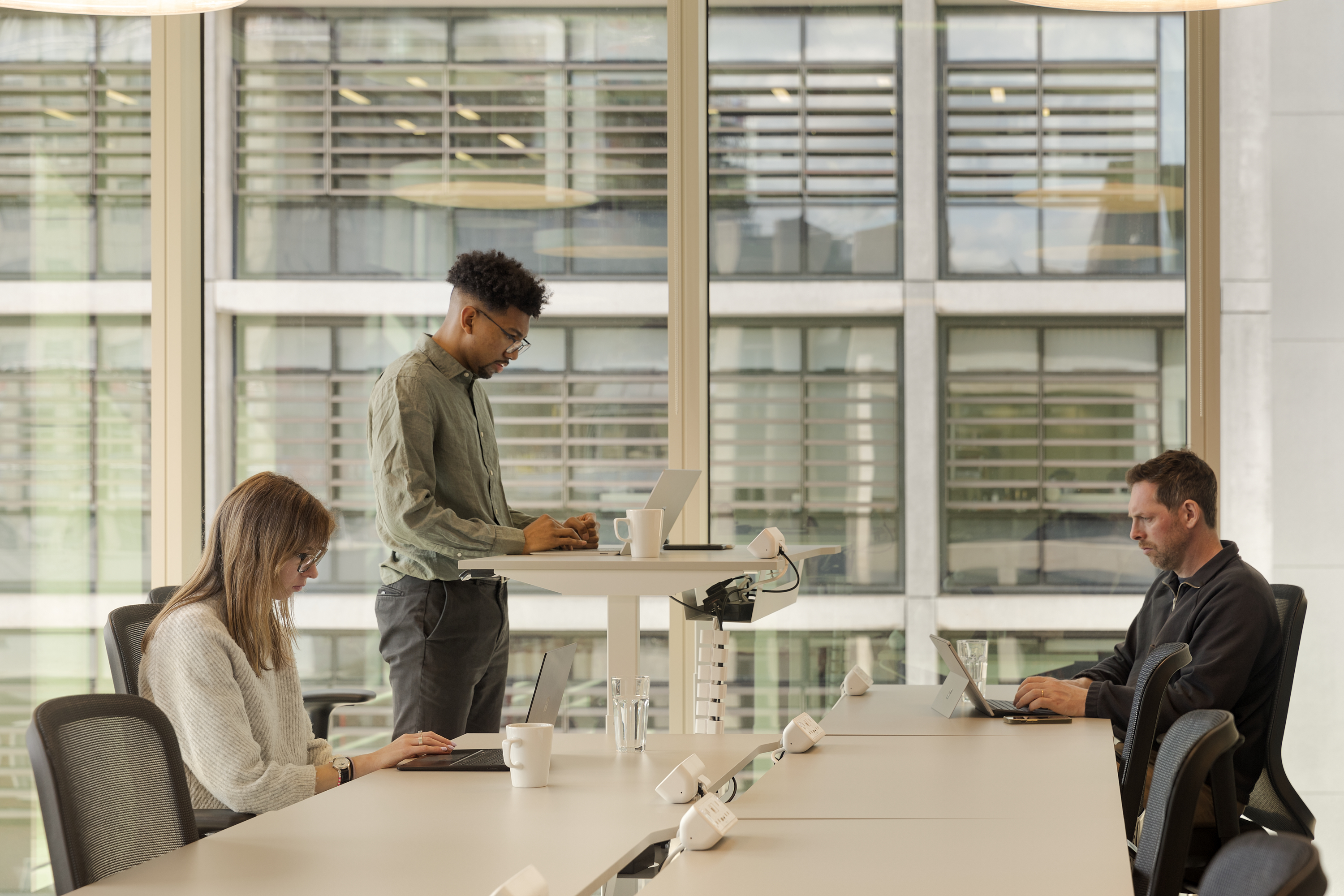 Shared workspace at MYO New Street Square featuring individuals working at ergonomic desks and standing-height workstations positioned beside floor-to-ceiling windows.