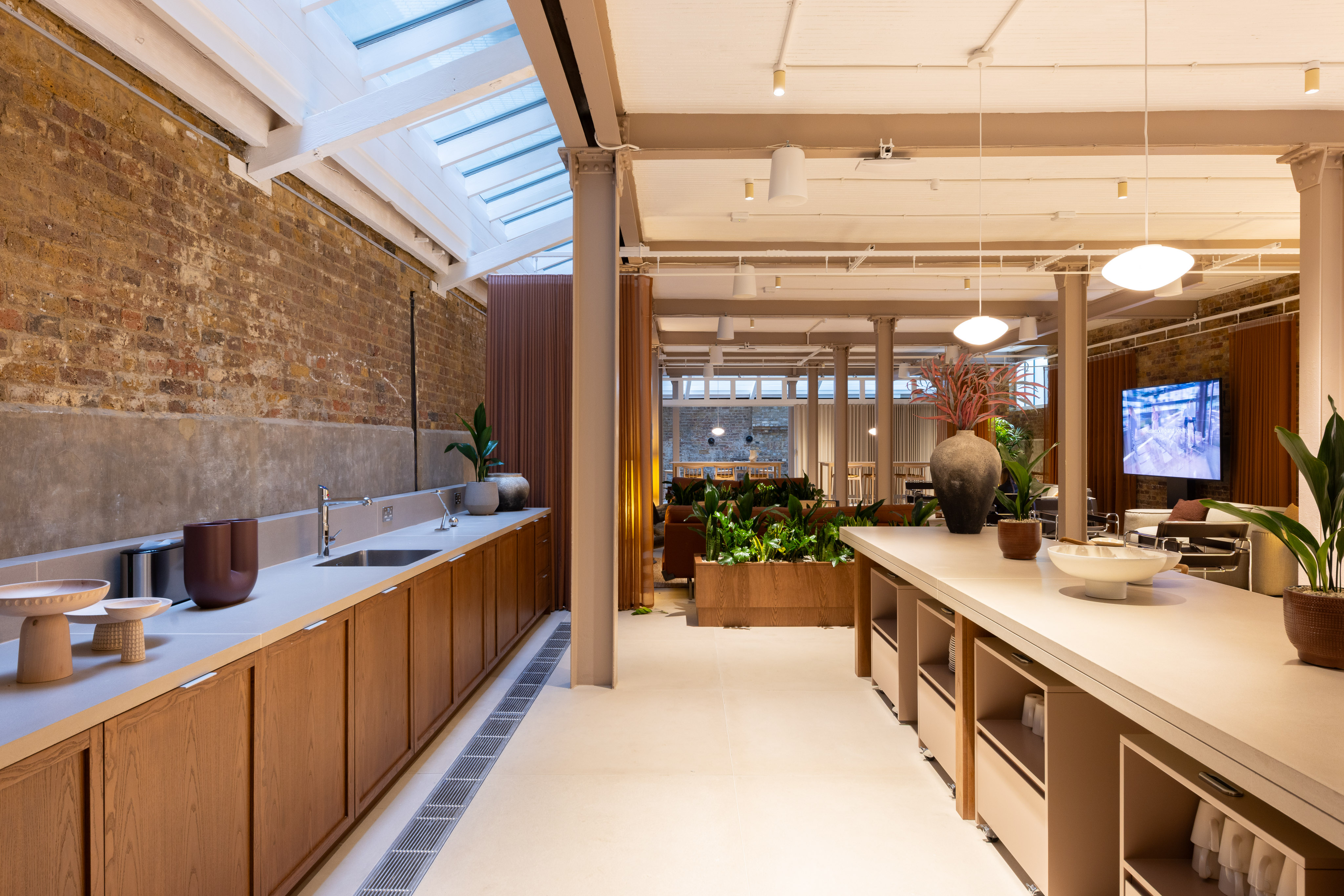 Open communal interior with a long kitchen counter, central island, lounge seating, indoor plants and exposed brick walls beneath skylights.