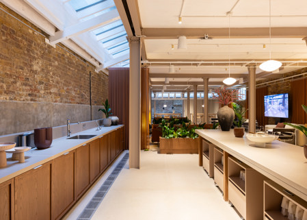 Open communal interior with a long kitchen counter, central island, lounge seating, indoor plants and exposed brick walls beneath skylights.