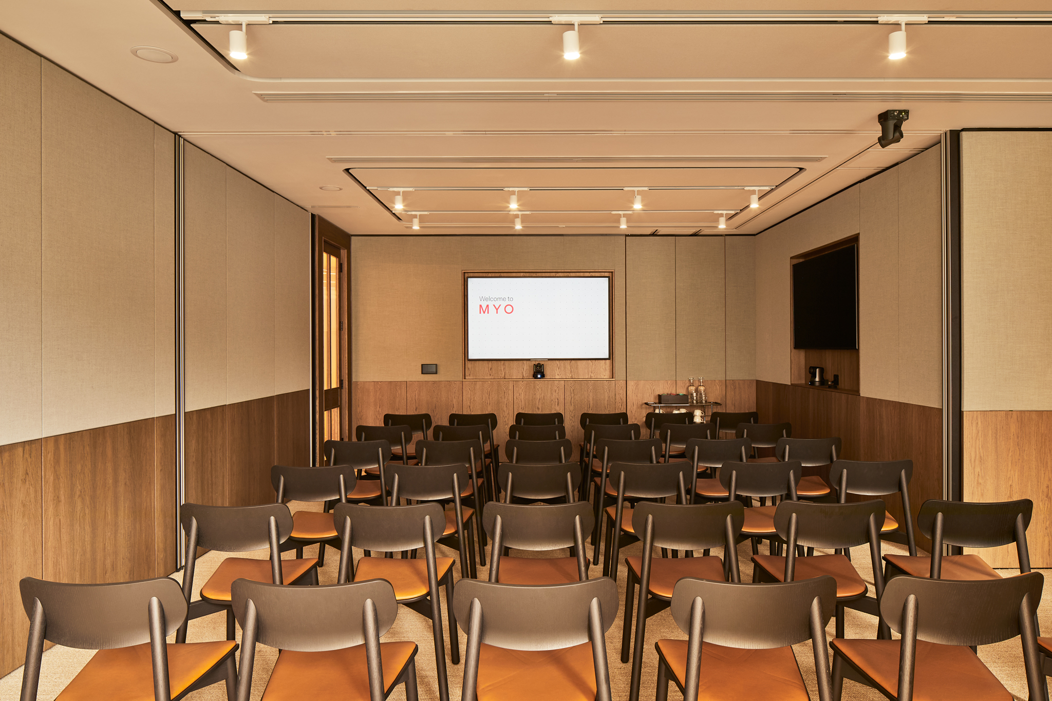 A contemporary meeting room arranged in a theatre-style layout with rows of chairs, dual presentation screens and modern wood-panelled walls.