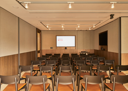 A contemporary meeting room arranged in a theatre-style layout with rows of chairs, dual presentation screens and modern wood-panelled walls.