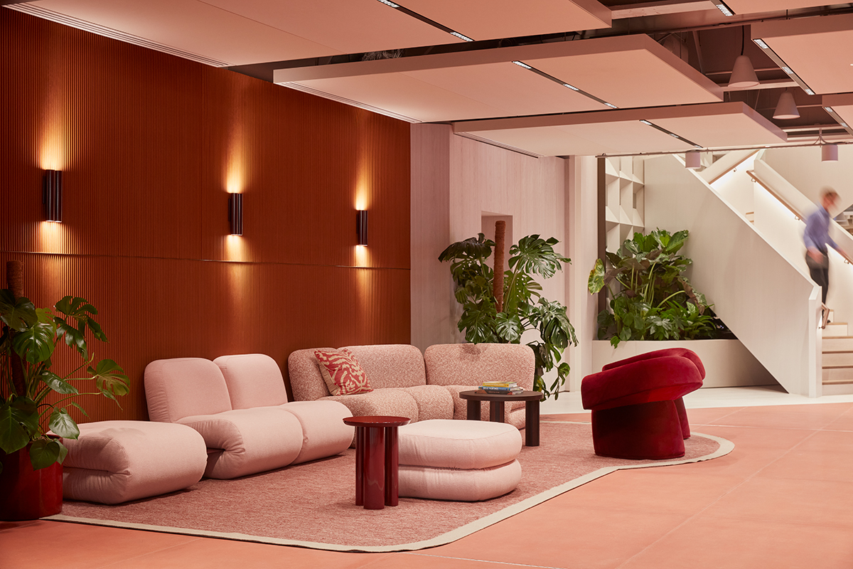 A modern lounge area featuring soft pink modular seating, a red accent chair, wooden wall panelling, indoor plants and an open staircase within a contemporary workspace.