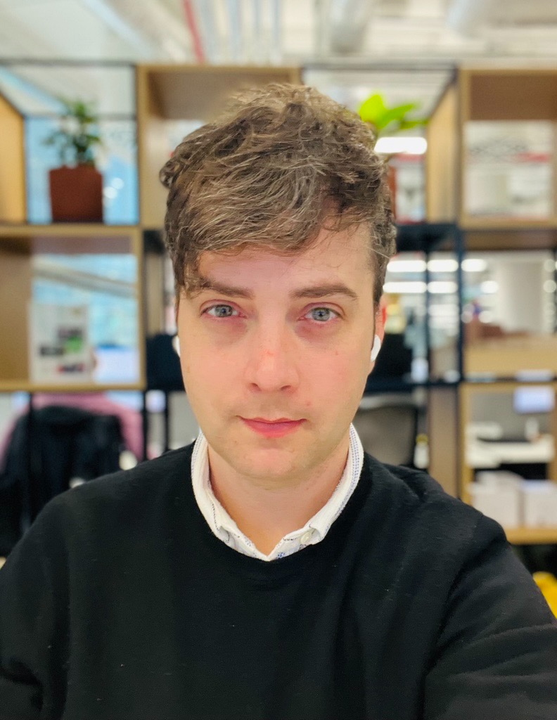Professional headshot of a man with short brown hair wearing a black jumper over a white collared shirt, photographed in front of open shelving with plants and workspace accessories in a MYO office setting.