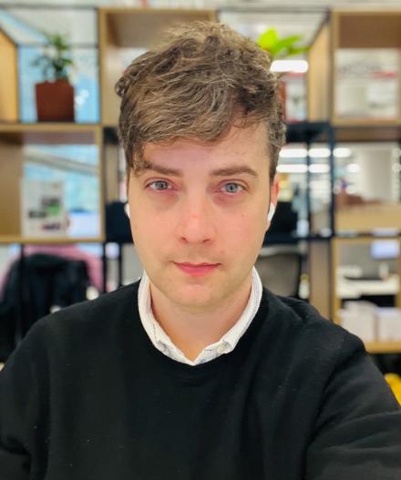 Professional headshot of a man with short brown hair wearing a black jumper over a white collared shirt, photographed in front of open shelving with plants and workspace accessories in a MYO office setting.