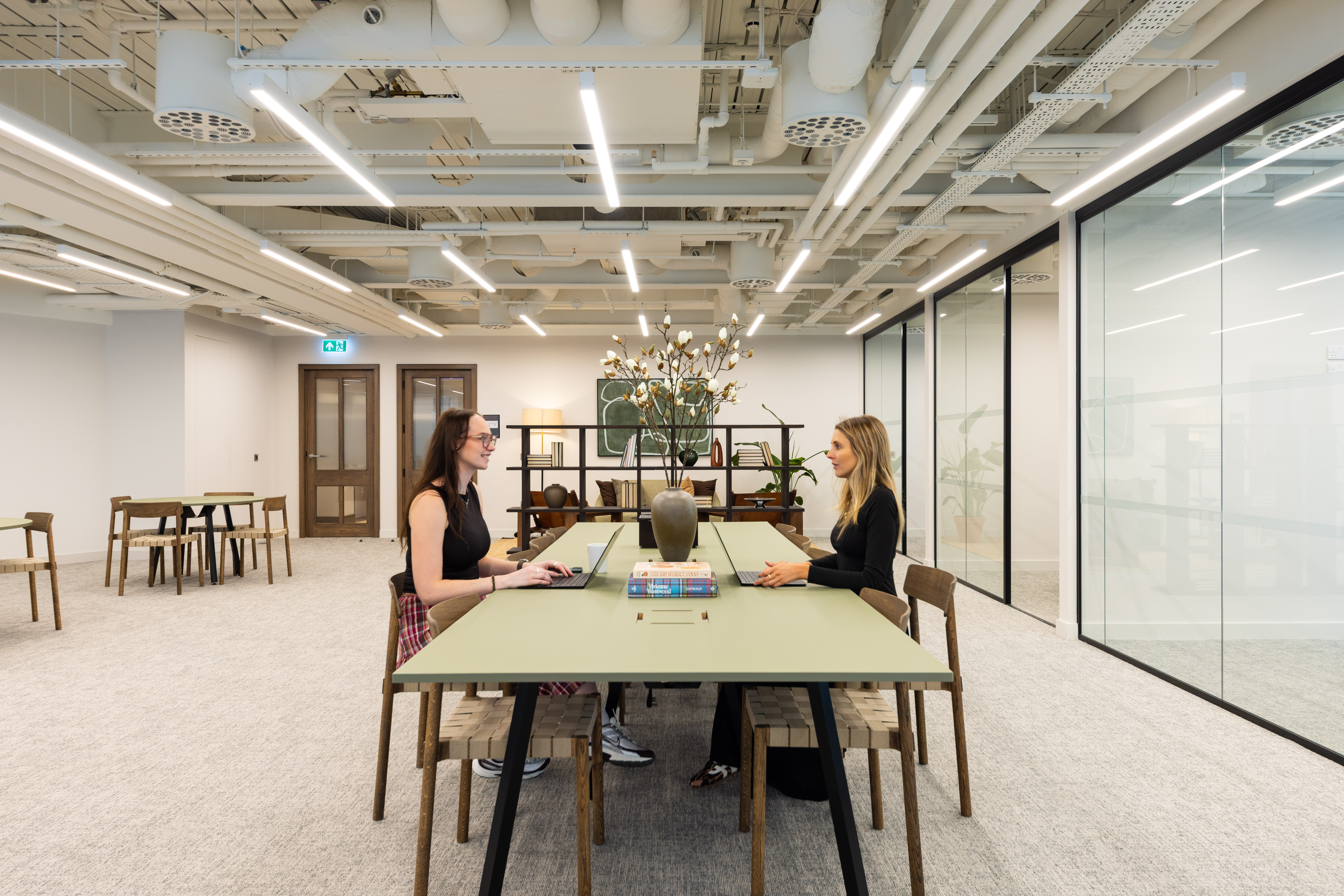 A contemporary open workspace with two people working at a shared table, surrounded by collaborative seating, glass meeting rooms and modern office interiors.