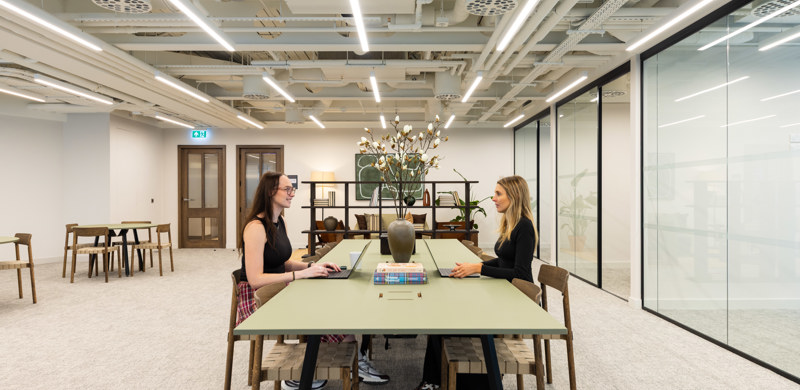 A contemporary open workspace with two people working at a shared table, surrounded by collaborative seating, glass meeting rooms and modern office interiors.