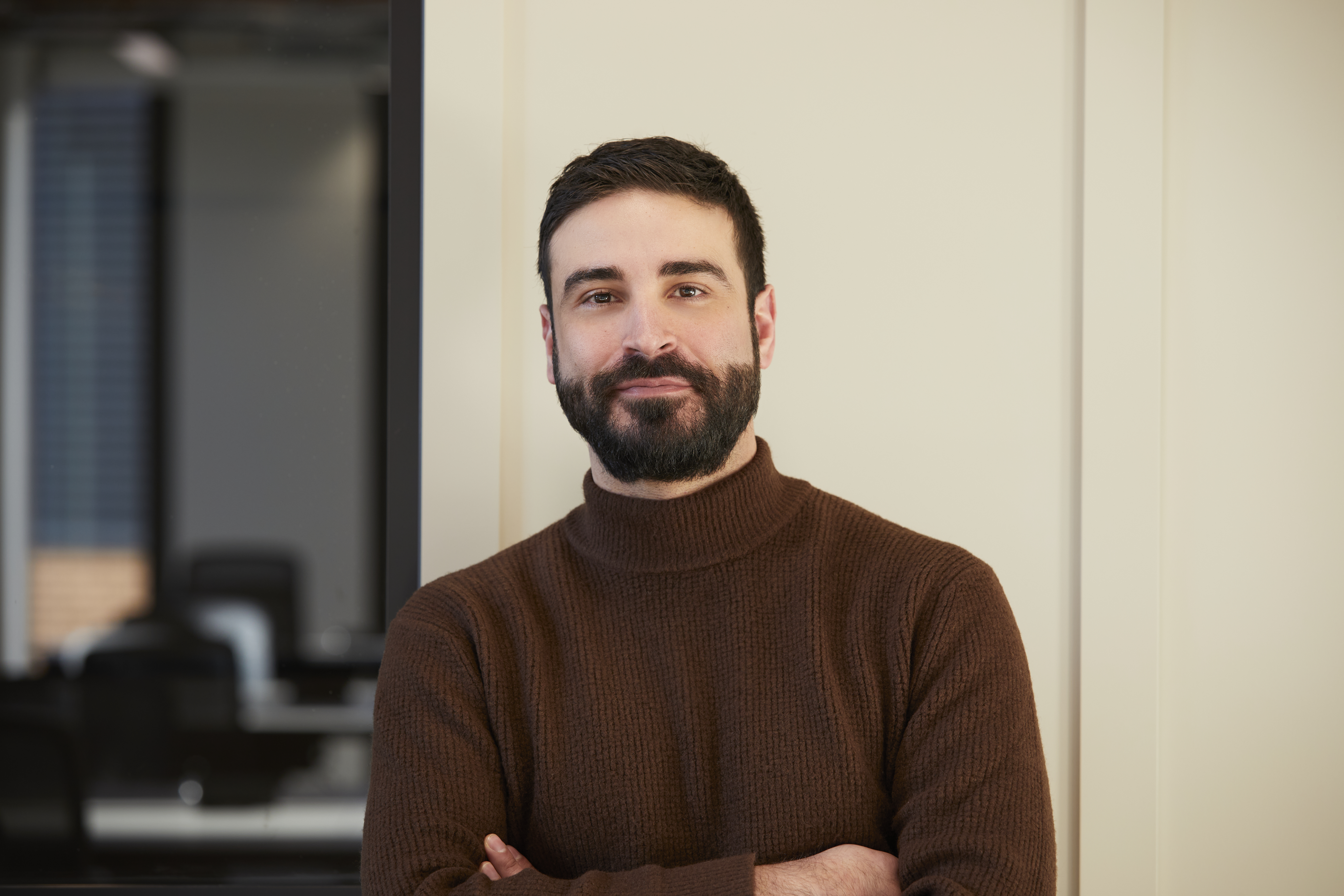 Professional headshot of a man with dark hair and a beard wearing a brown roll-neck jumper, standing with folded arms against a neutral-toned backdrop within a contemporary MYO workspace.