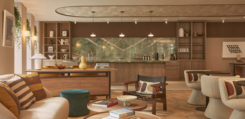 A contemporary lounge and kitchen area featuring comfortable seating, wooden coffee tables, a communal kitchen, open shelving and warm interior finishes.