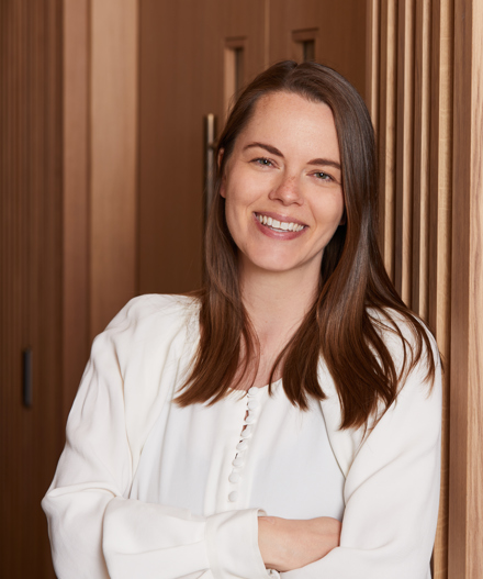 Professional headshot of a smiling woman with shoulder-length brown hair wearing a white blouse, standing beside light oak slatted wall panelling within a MYO workspace.