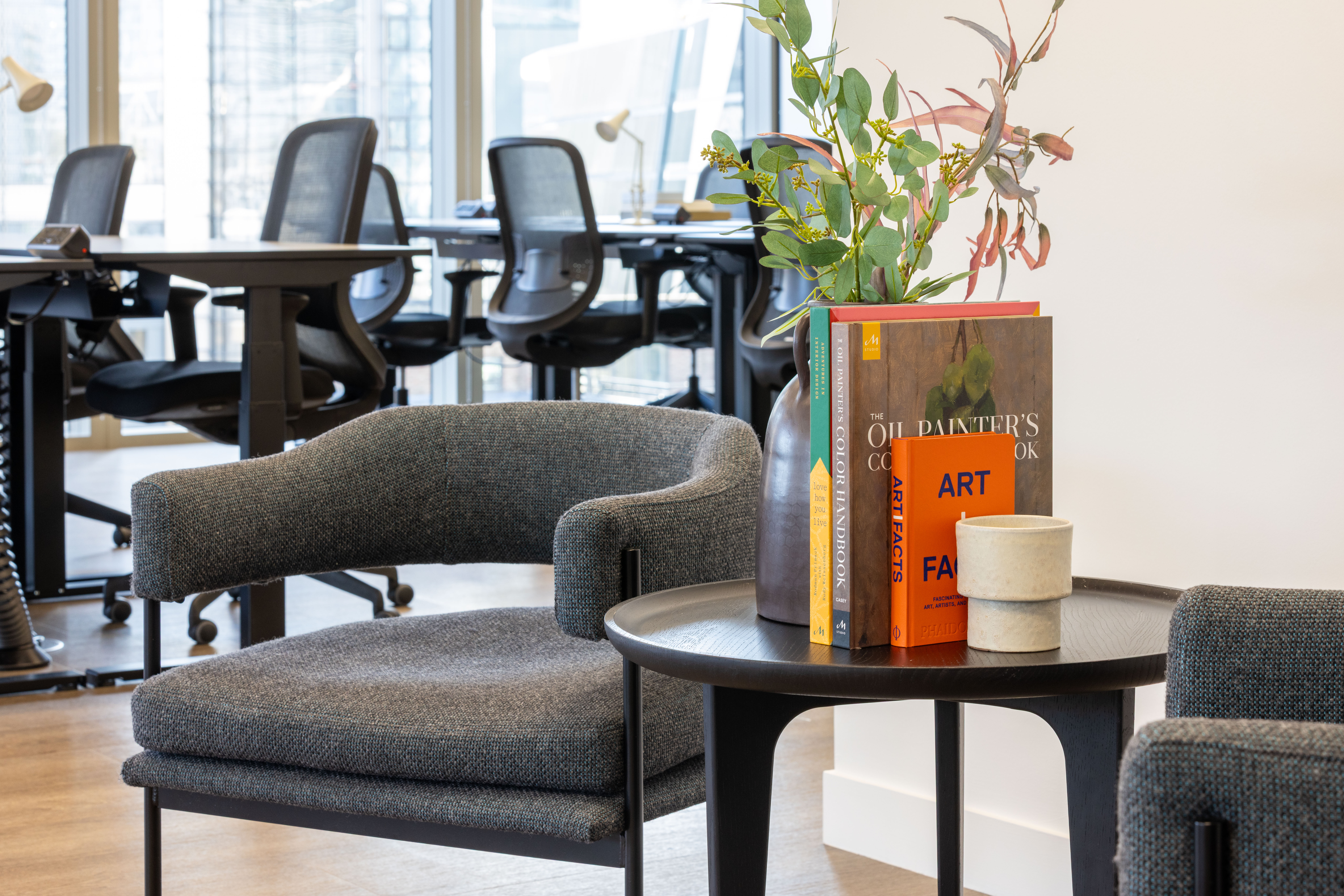 Comfortable lounge seating area at MYO Liverpool Street featuring textured grey armchairs, curated books, fresh florals and open-plan workspace in the background.