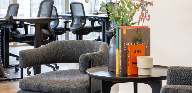 Comfortable lounge seating area at MYO Liverpool Street featuring textured grey armchairs, curated books, fresh florals and open-plan workspace in the background.