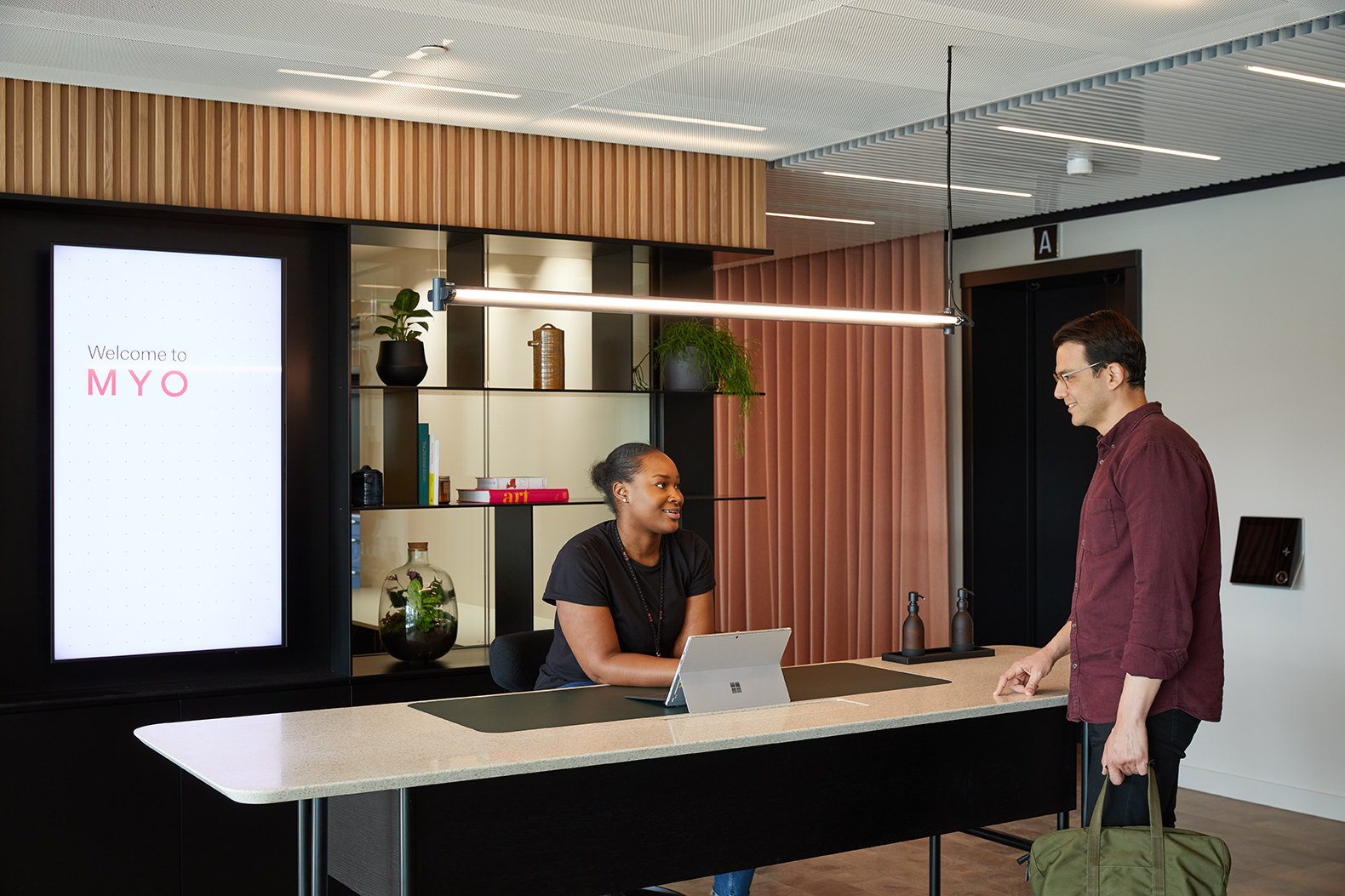 Reception area at MYO Liverpool Street where a member of the Experience Team welcomes a visitor at a contemporary desk framed by blush-toned panelling and curated shelving.