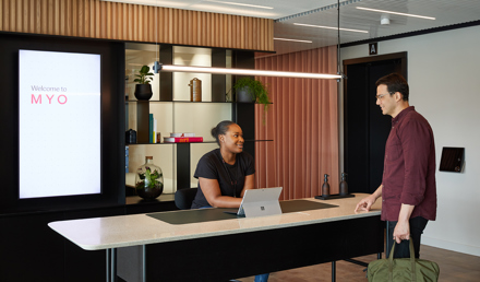 Reception area at MYO Liverpool Street where a member of the Experience Team welcomes a visitor at a contemporary desk framed by blush-toned panelling and curated shelving.