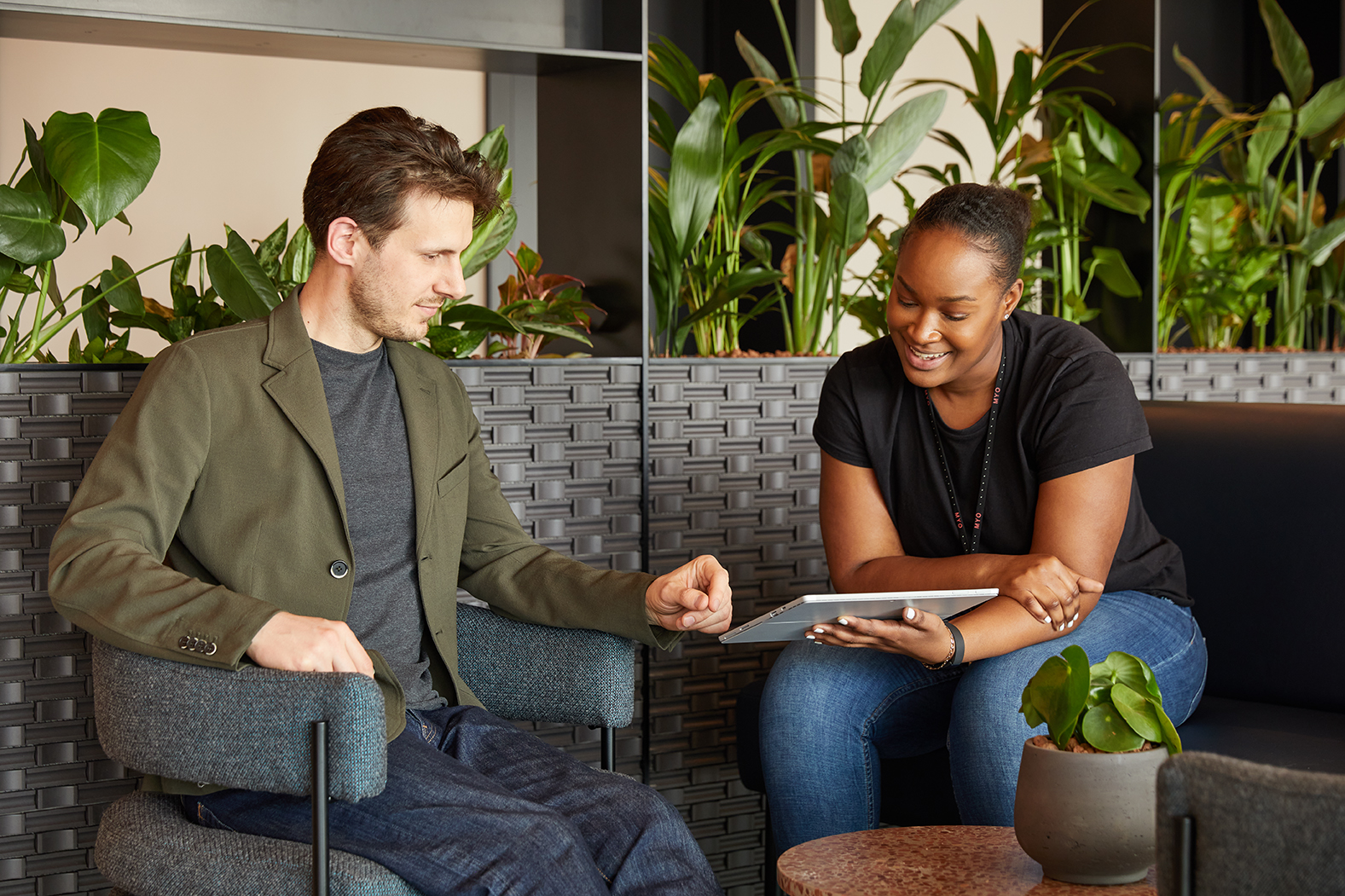 MYO team member supporting a colleague with a tablet in a welcoming lounge area surrounded by greenery.