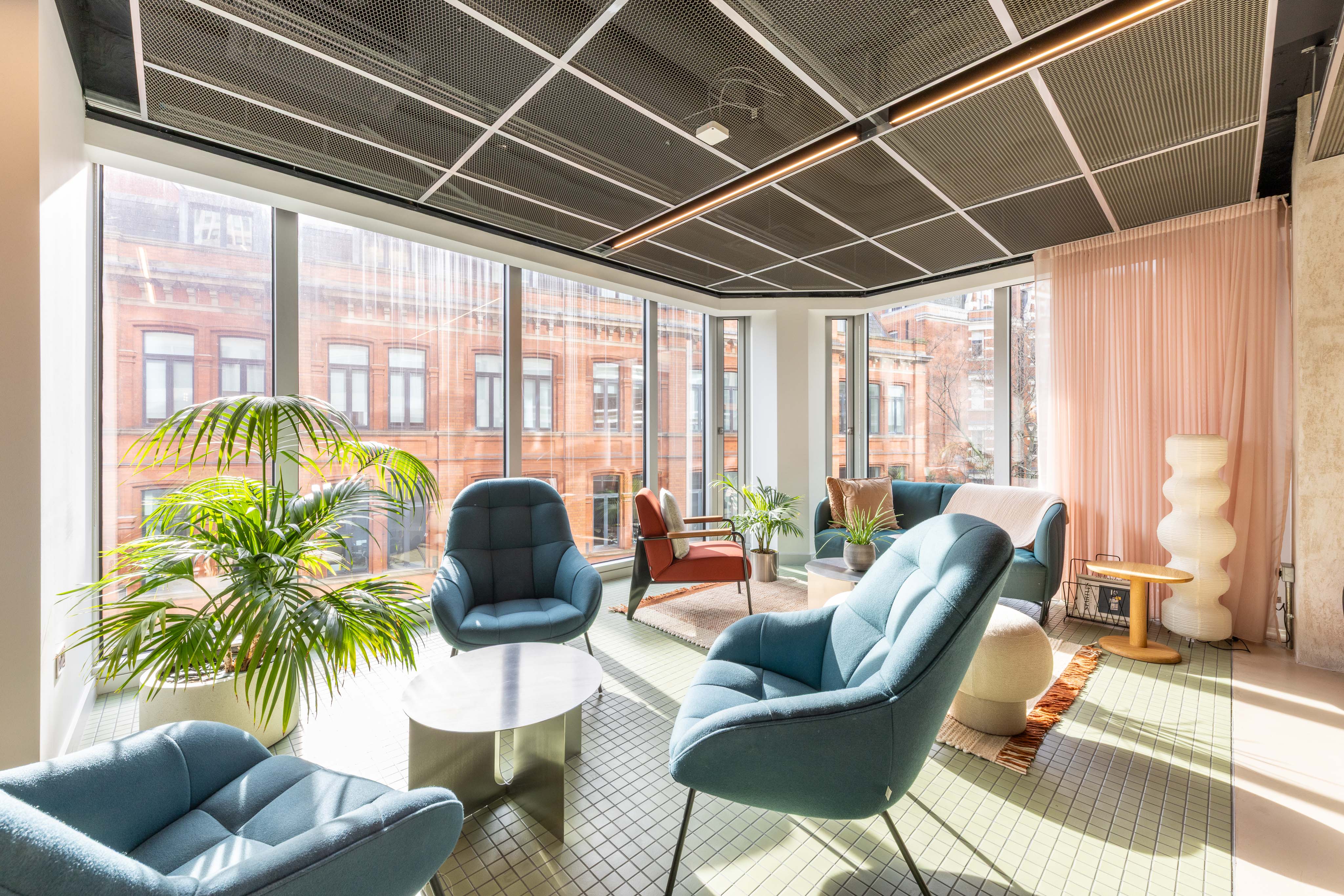 A contemporary lounge area with blue armchairs, sofas, coffee tables, indoor plants and floor-to-ceiling windows overlooking the city.