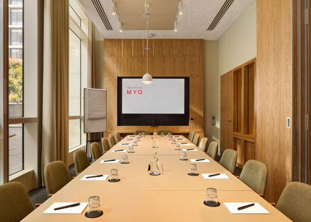 A contemporary boardroom featuring a long conference table, upholstered chairs, a presentation screen, flip chart and floor-to-ceiling windows.