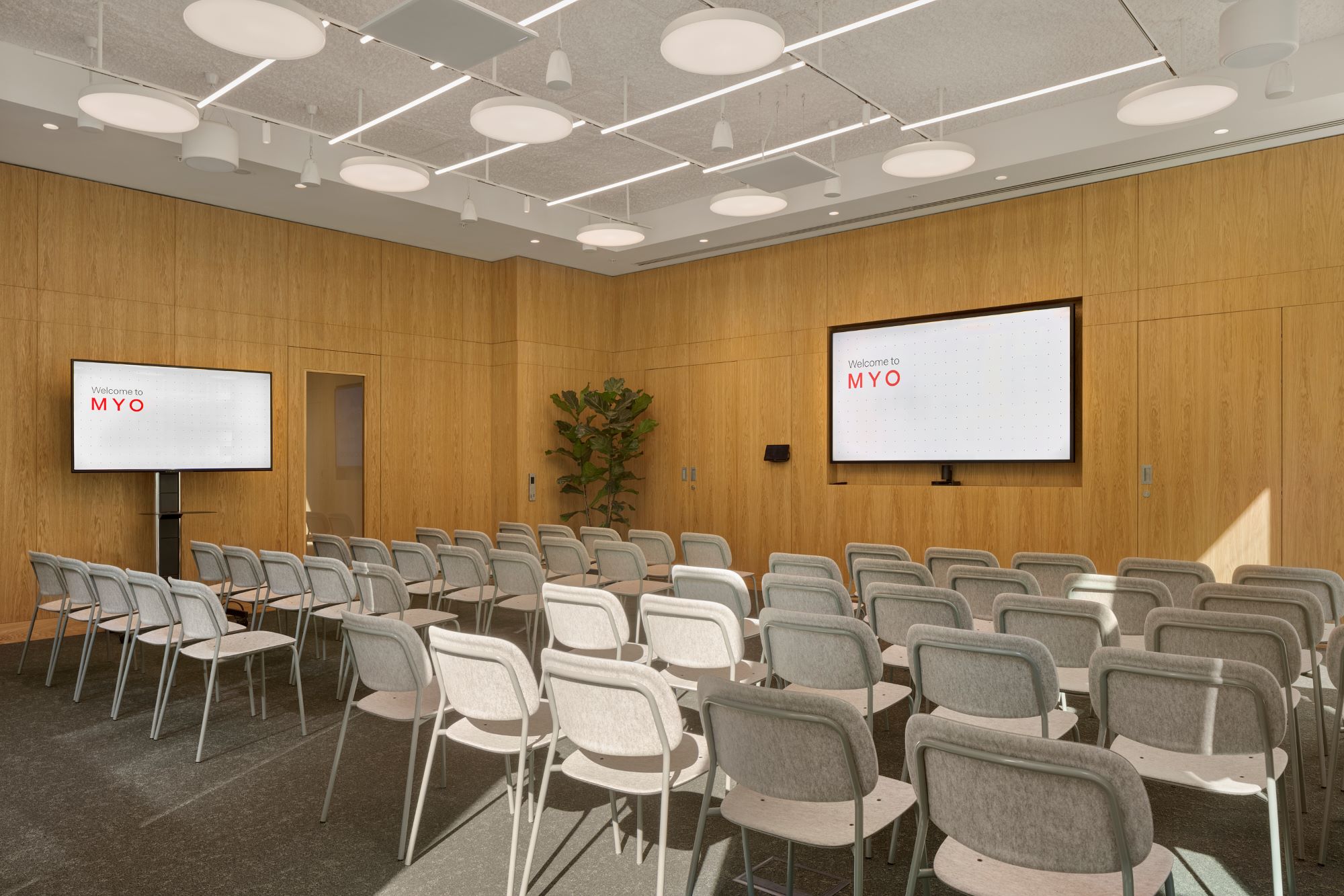 A contemporary conference room with rows of upholstered chairs, two large presentation screens, timber wall panelling and modern lighting.