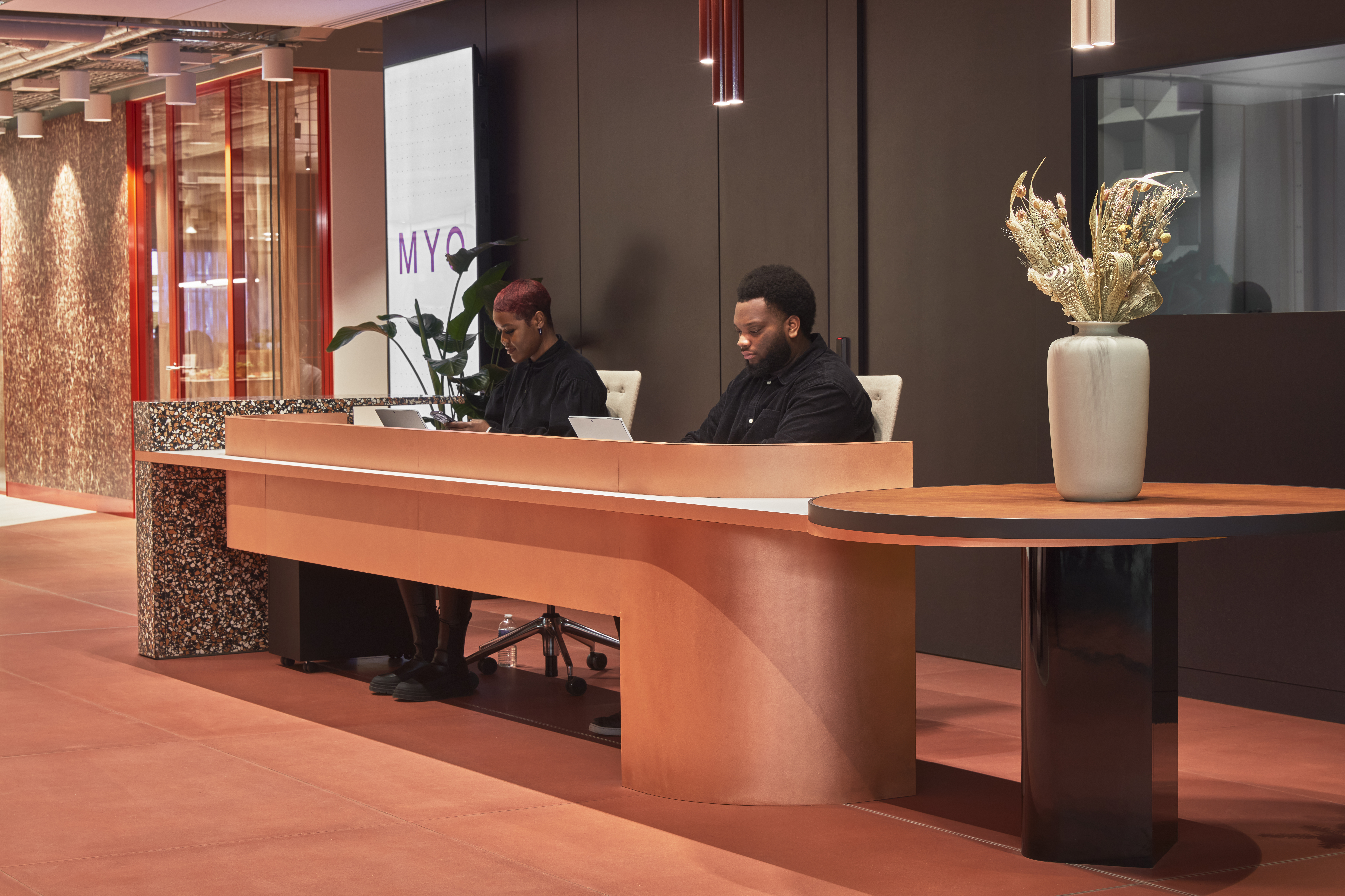 Reception area with two staff members seated behind a curved wooden desk, with dark wall panelling, pendant lights and a vase on a side table.