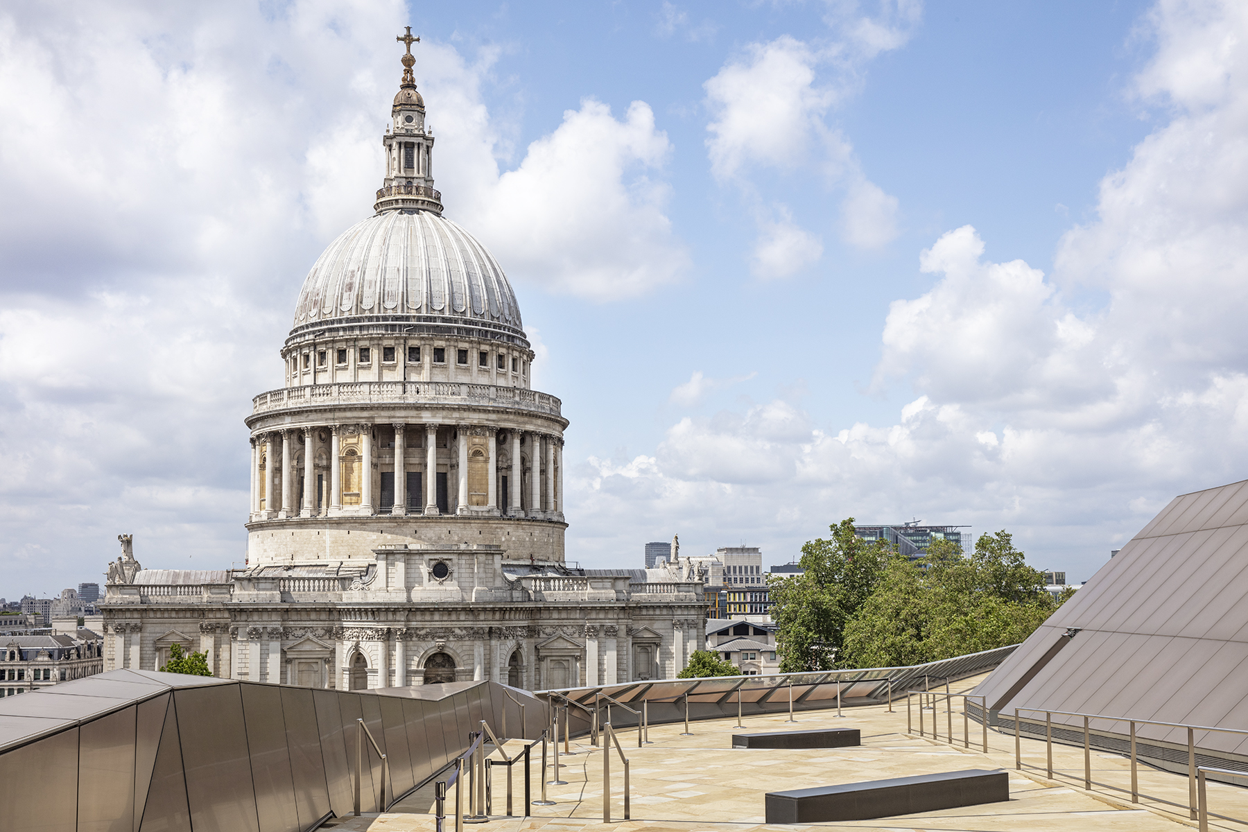 Rooftop terrace with stone paving and railings overlooking St Paul’s Cathedral under a blue sky with clouds.