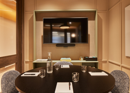A contemporary meeting room featuring a round conference table, upholstered chairs, a wall-mounted presentation screen, integrated video conferencing equipment and warm timber finishes.