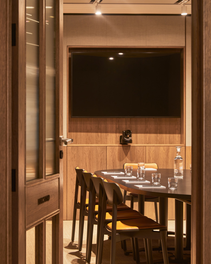 A modern meeting room with a conference table, chairs, video conferencing equipment and a wall-mounted display, viewed through an open doorway.