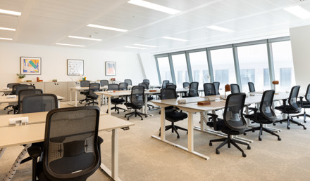 Bright open-plan office with rows of white desks, black mesh office chairs and large slanted windows along one side.