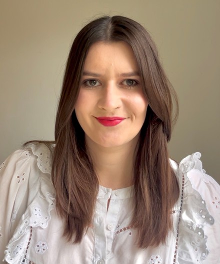 Professional headshot of a woman with shoulder-length dark brown hair wearing a white embroidered blouse with broderie detailing, photographed against a soft neutral background.
