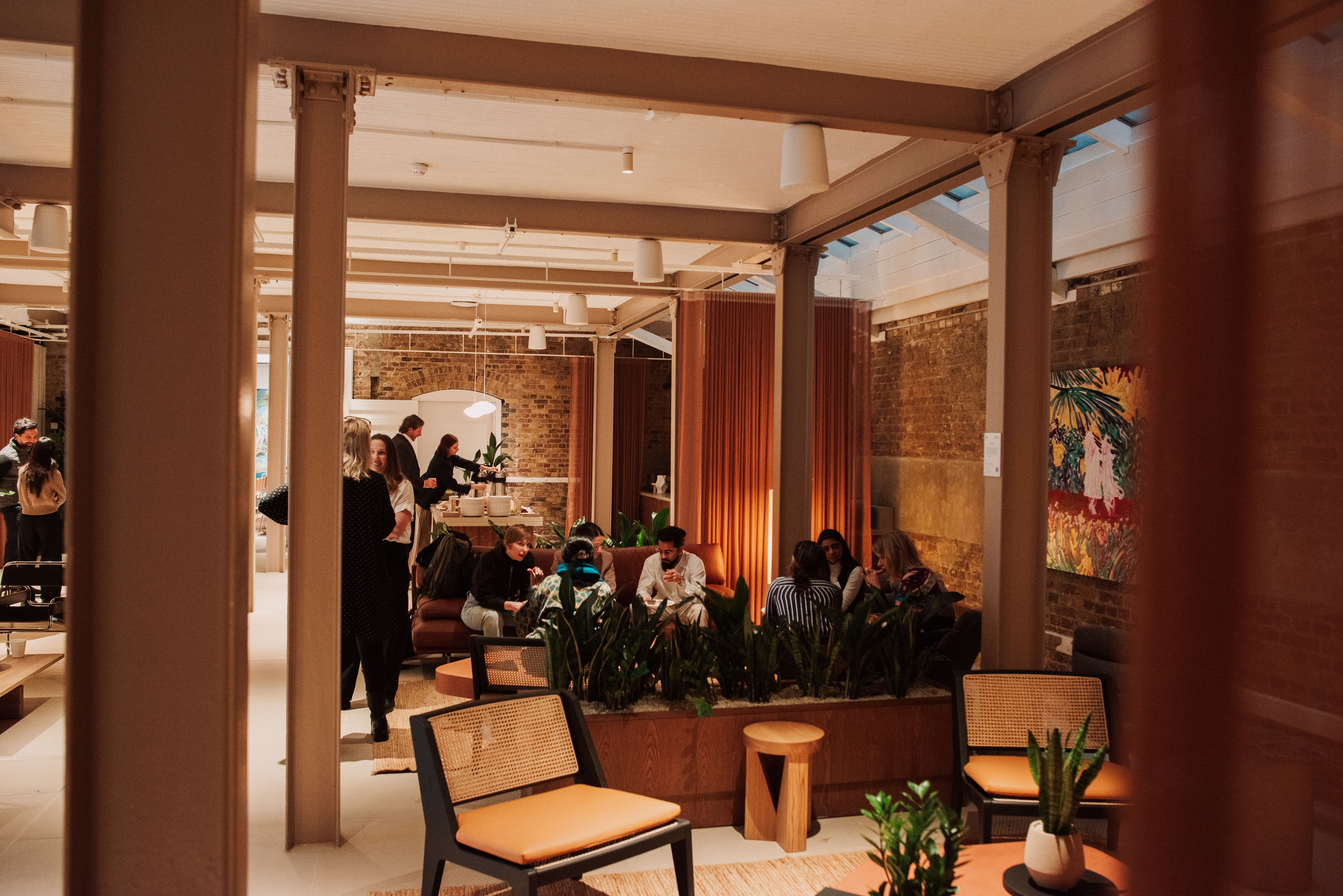 A contemporary communal lounge with people socialising, comfortable seating, indoor planting and exposed brick walls in a modern workspace.