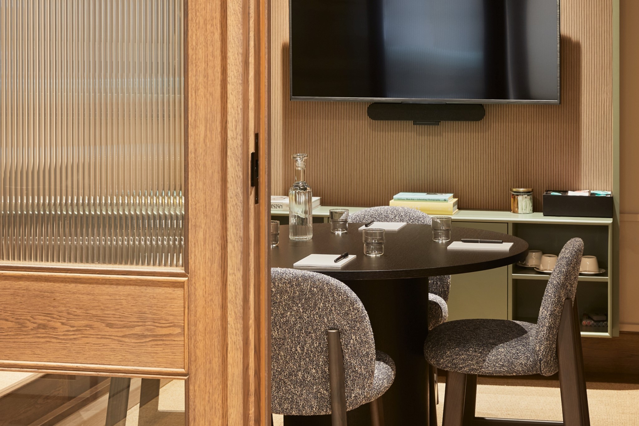 A contemporary meeting room viewed through a glazed timber door, featuring a round meeting table, upholstered chairs, a wall-mounted presentation screen and integrated video conferencing equipment.