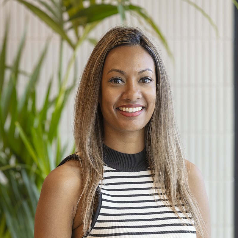 Professional headshot of a smiling woman with long highlighted hair wearing a black and white striped sleeveless top, standing in front of a softly blurred background with green planting and textured wall panels.