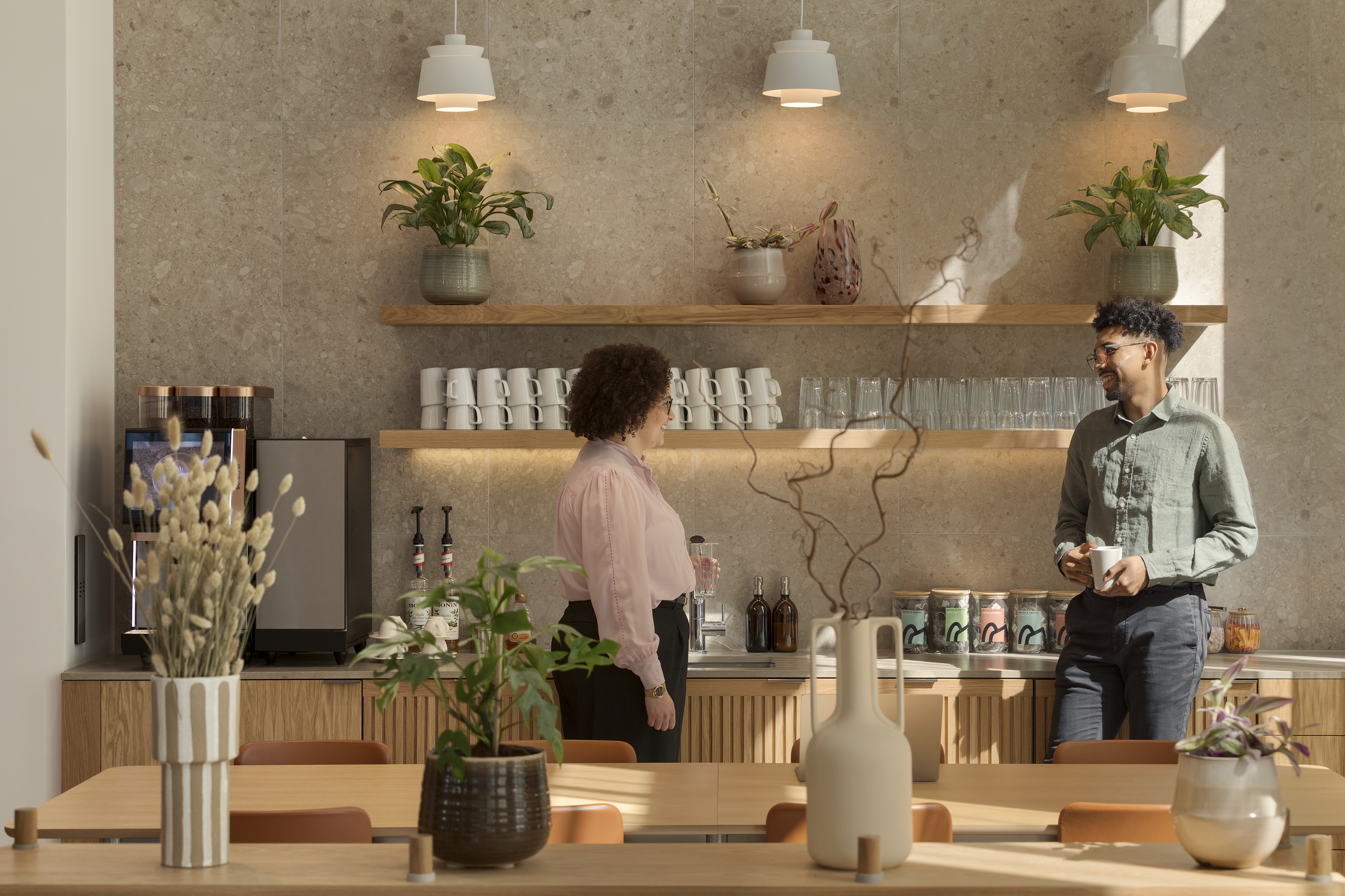 A contemporary communal kitchen featuring a coffee station, open shelving, indoor plants and two people chatting in a modern workspace.