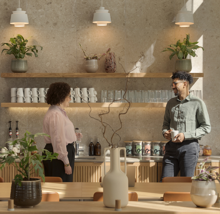 A contemporary communal kitchen featuring a coffee station, open shelving, indoor plants and two people chatting in a modern workspace.