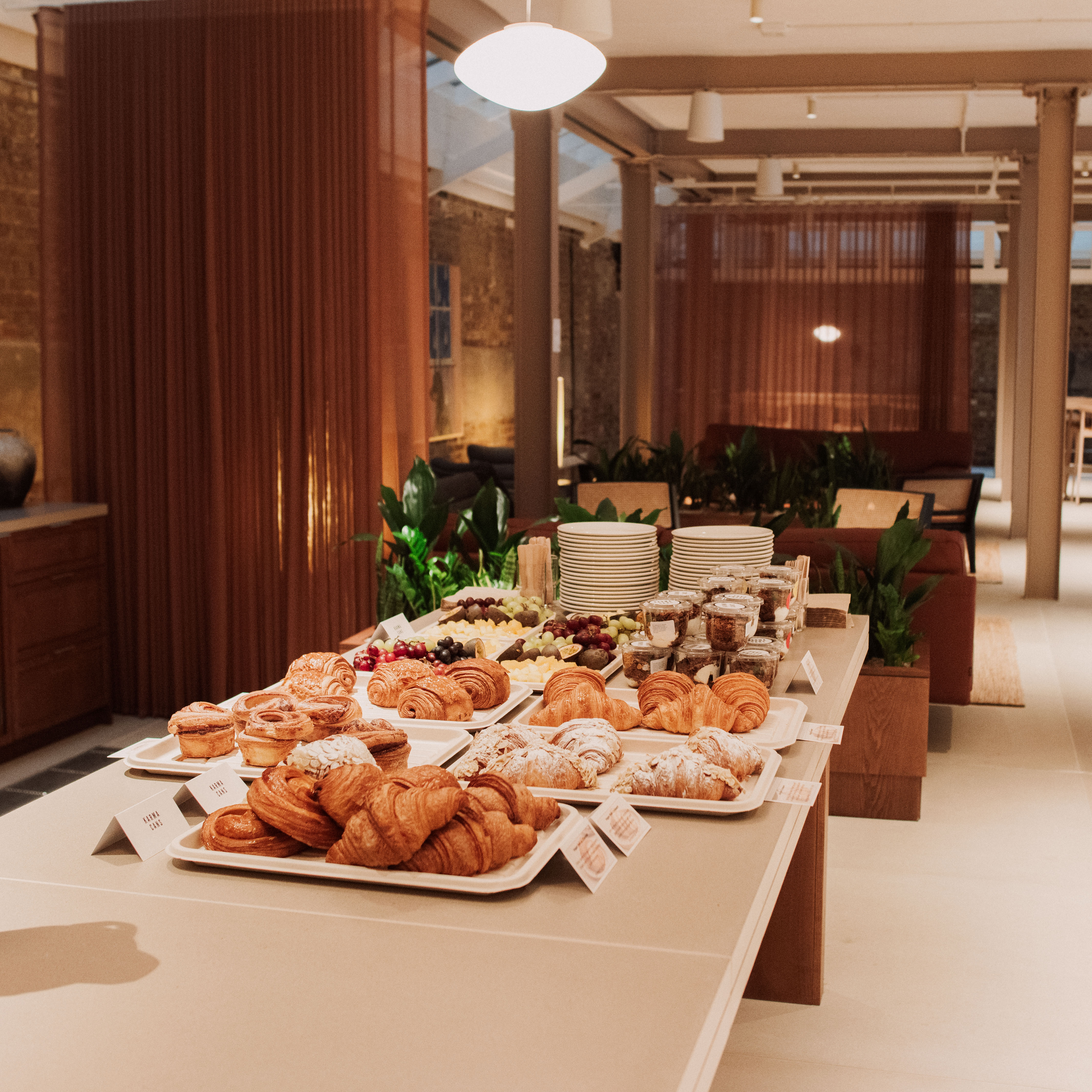 Buffet table with trays of pastries, fruit, yoghurt pots and stacked plates in a lounge-style event space.