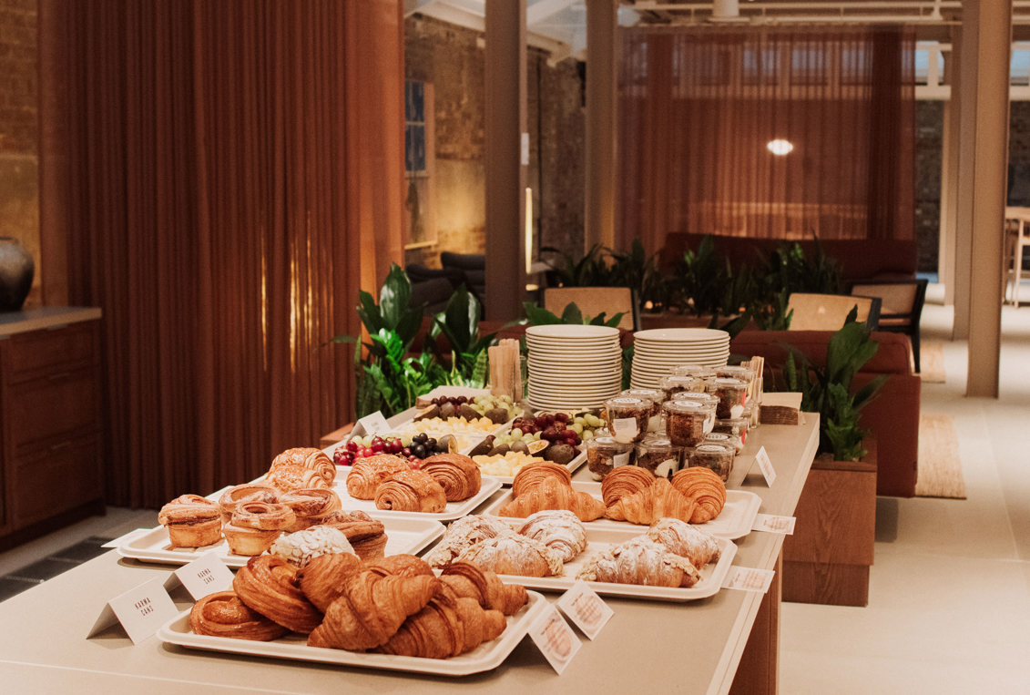 Buffet table with trays of pastries, fruit, yoghurt pots and stacked plates in a lounge-style event space.
