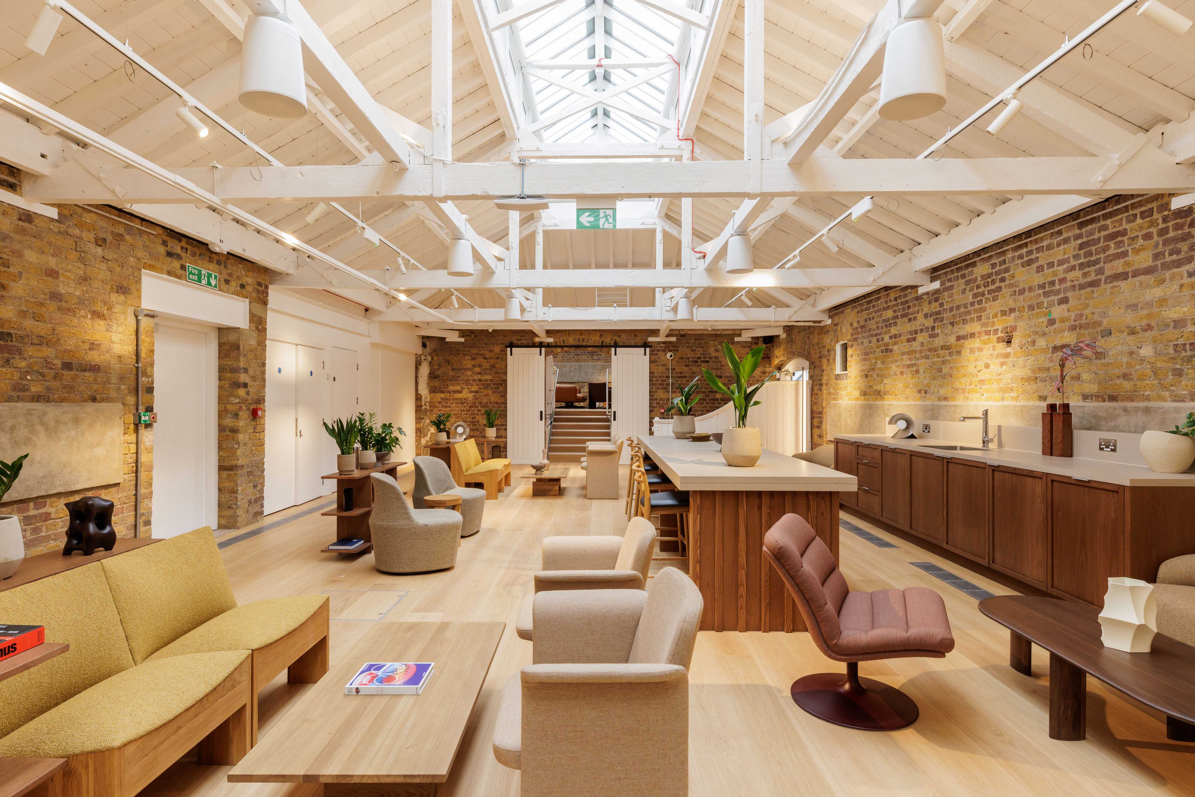 Open lounge-style workspace with sofas, armchairs, a central island counter, brick walls and a high white ceiling with skylights.