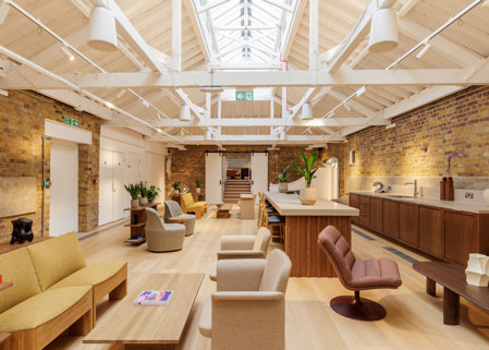 Open lounge-style workspace with sofas, armchairs, a central island counter, brick walls and a high white ceiling with skylights.