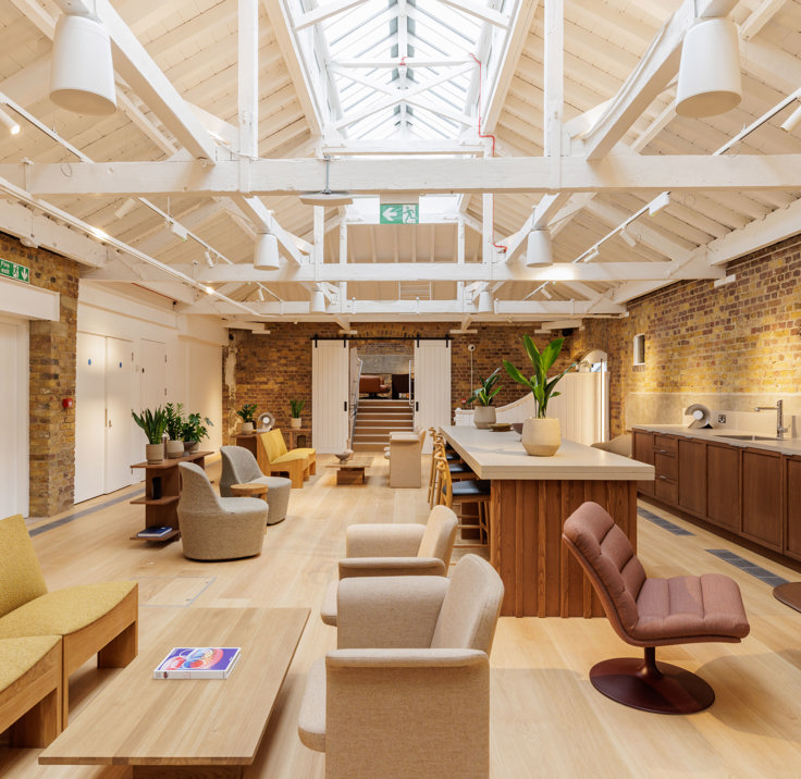 Open lounge-style workspace with sofas, armchairs, a central island counter, brick walls and a high white ceiling with skylights.