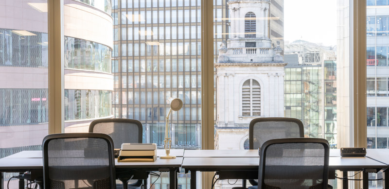 Dedicated workstations at MYO Liverpool Street with floor-to-ceiling windows overlooking the surrounding city skyline and historic architecture.
