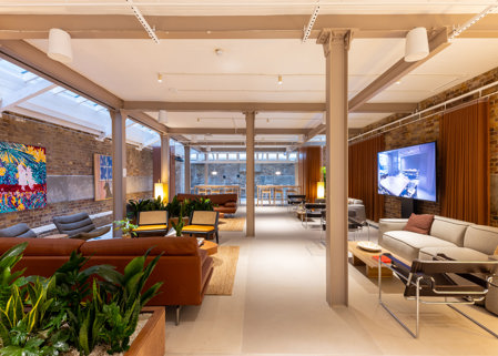 Spacious lounge-style workspace with sofas, armchairs, indoor plants, exposed brick walls, colourful artwork and a wall-mounted screen.