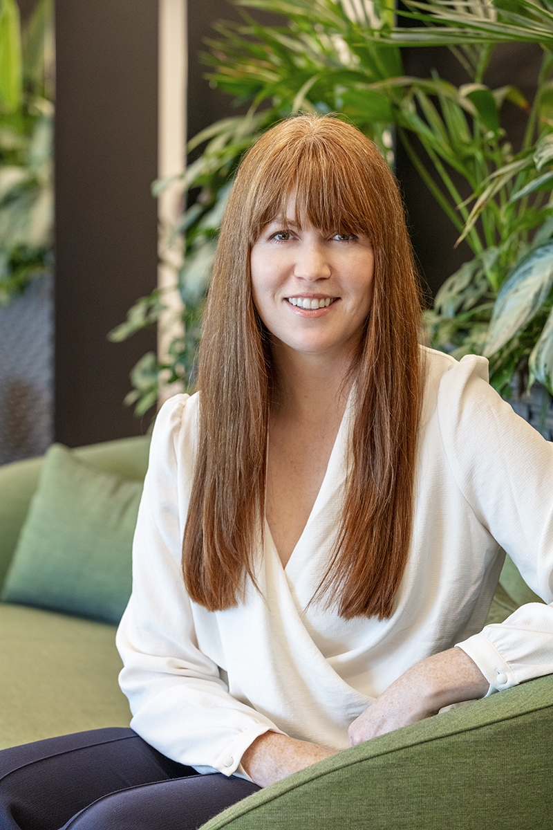 Professional headshot of a smiling woman with long auburn hair and a fringe, wearing a white blouse and seated on a green upholstered sofa surrounded by indoor planting within a MYO workspace.
