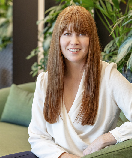Professional headshot of a smiling woman with long auburn hair and a fringe, wearing a white blouse and seated on a green upholstered sofa surrounded by indoor planting within a MYO workspace.