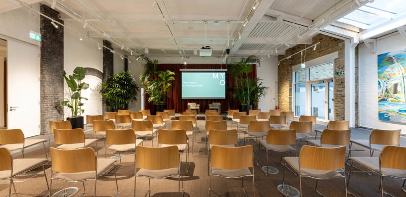 A contemporary event space with rows of chairs facing a presentation screen, surrounded by indoor plants, exposed brick walls and modern lighting.