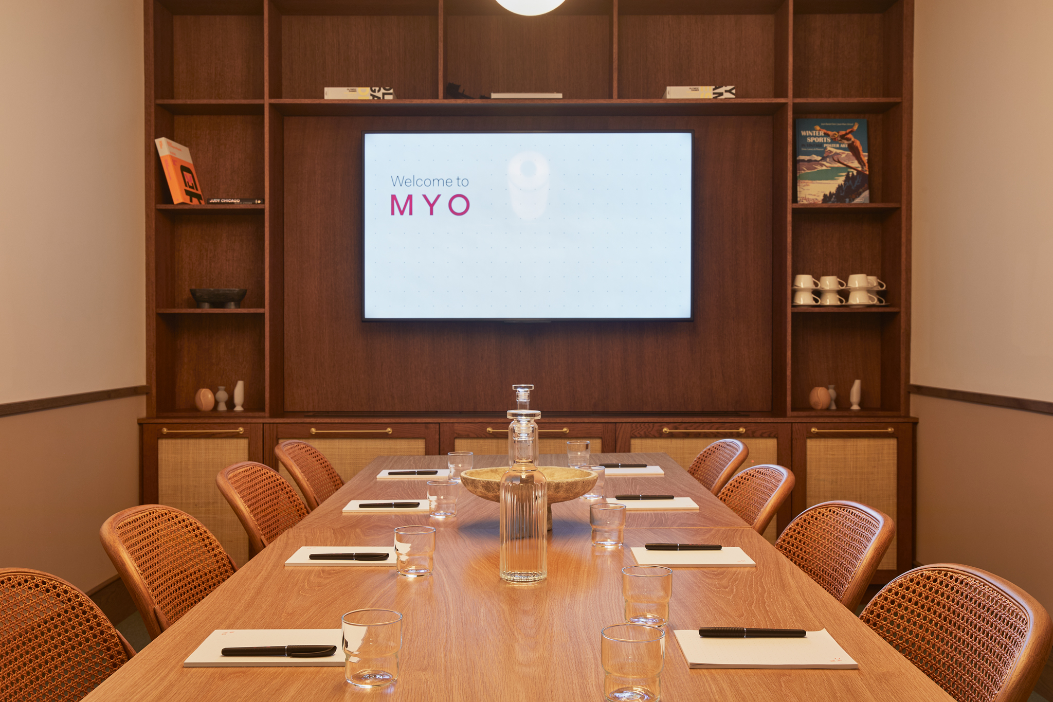 A contemporary meeting room featuring a wooden conference table, woven chairs, a built-in presentation screen and timber shelving with decorative accessories.