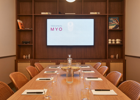 A contemporary meeting room featuring a wooden conference table, woven chairs, a built-in presentation screen and timber shelving with decorative accessories.