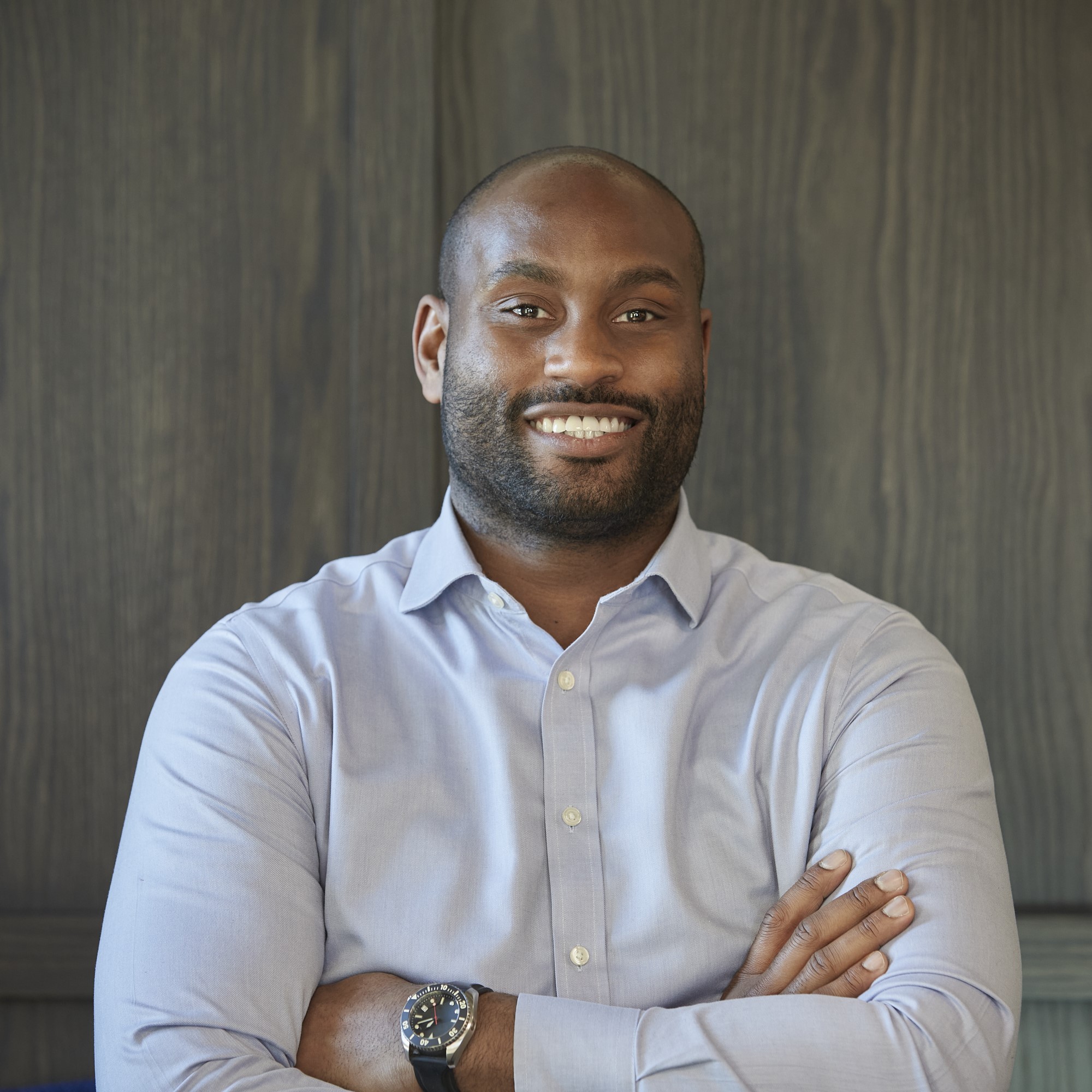 Professional headshot of a smiling man with a shaved head and trimmed beard, wearing a light blue button-down shirt and wristwatch, standing with folded arms against a dark wood-panelled backdrop.