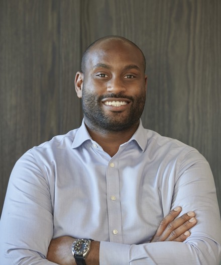 Professional headshot of a smiling man with a shaved head and trimmed beard, wearing a light blue button-down shirt and wristwatch, standing with folded arms against a dark wood-panelled backdrop.
